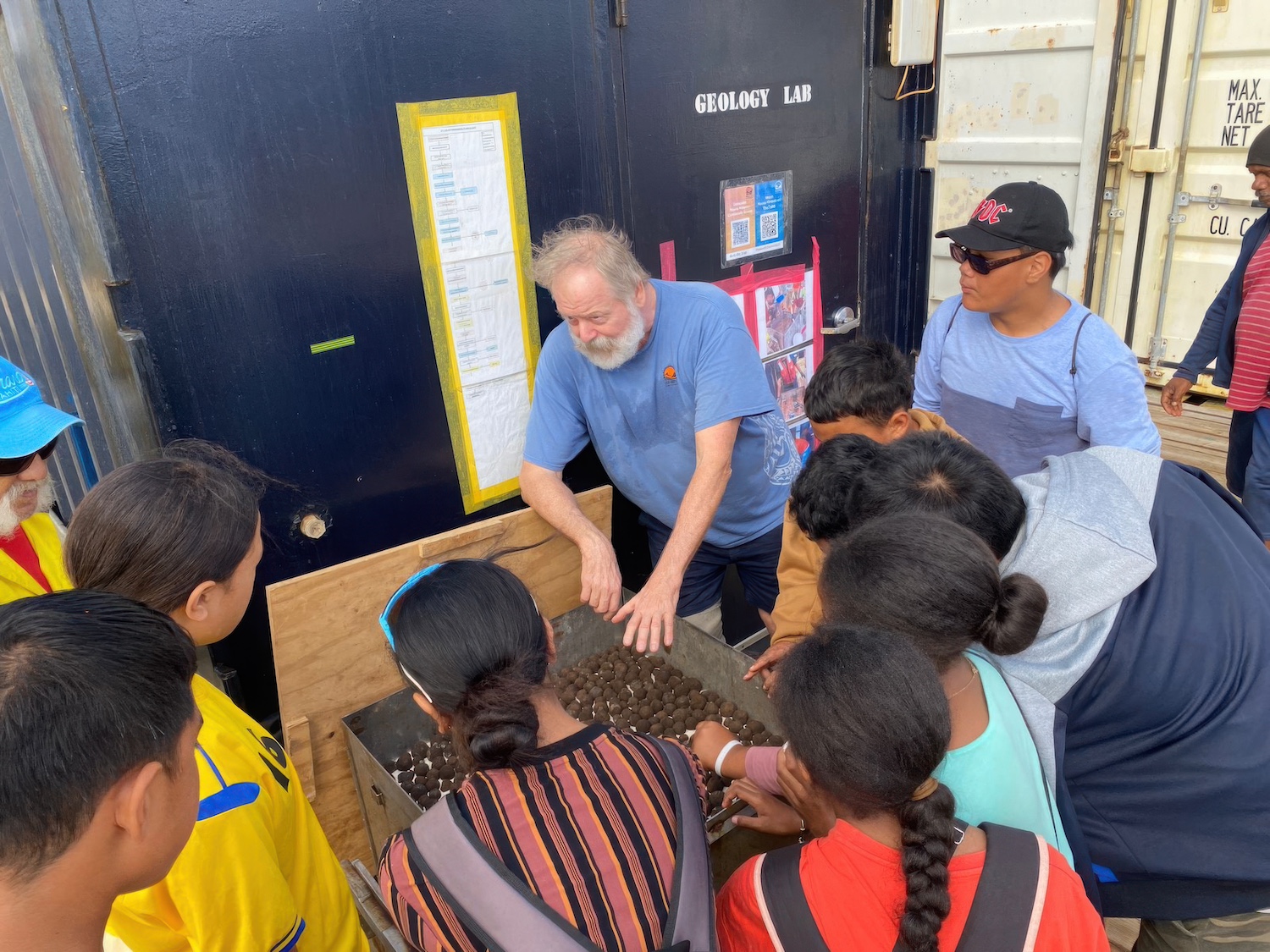 The polymetallic nodules that can be found on the ocean floor, resembling small brown spheres of rock, are displayed in a box while John McIntyre talks about them with Cook Islanders — deep-sea mining